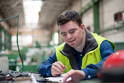 Zwischen Förderung und Ausgrenzung: Die Debatte um Behindertenwerkstätten Ein junger Mann mit Downsyndrom sitzt in einer großen Werkshalle an einem Tisch und notiert etwas auf einem Zettel. Er trät eine neonfarbene Warnweste.