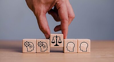 Diskriminierung durch Künstliche Intelligenz: Ein Einblick in die systemischen Risiken Eine Hand hebt den mittleren von fünf Holzwürfeln leicht nach oben an. Auf ihm ist eine Waage erkennbar.