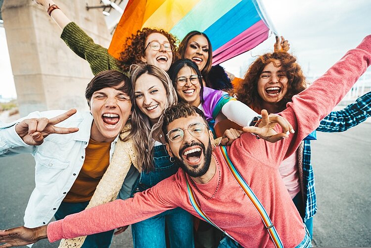 Pride Month: Ein Fest der Freude, der Triumphe und des Leids der LGBTQIA+ Community Eine Gruppe fröhlicher, junger Menschen aus der LGBQTI+-Community lacht in die Kamera. Dabei machen einige von ihnen ein das Peace-Zeichen, eine Person hält eine Regenbogenflagge.
