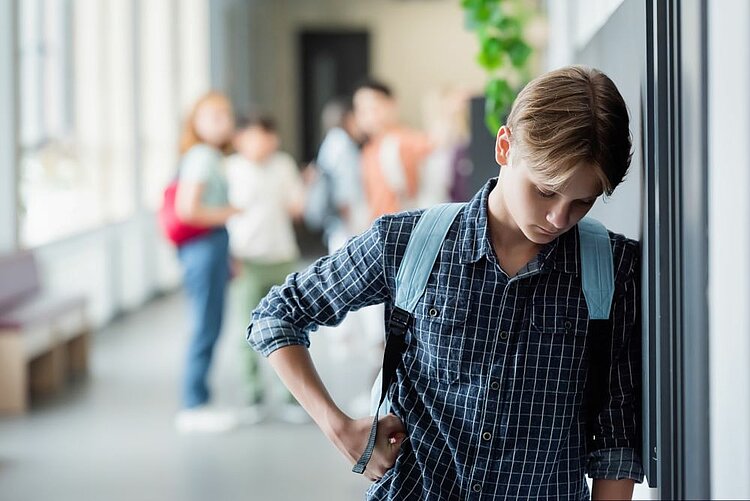 Ein Jugendlicher mit einem Rucksack steht allein in einem Schulkorridor, lehnt sich gegen eine Wand und lässt den Kopf traurig hängen. Im Hintergrund stehen andere Schülerinnen und Schüler zusammen und schauen zu ihm rüber und reden scheinbar über ihn. Das Bild vermittelt eine bedrückende Atmosphäre.