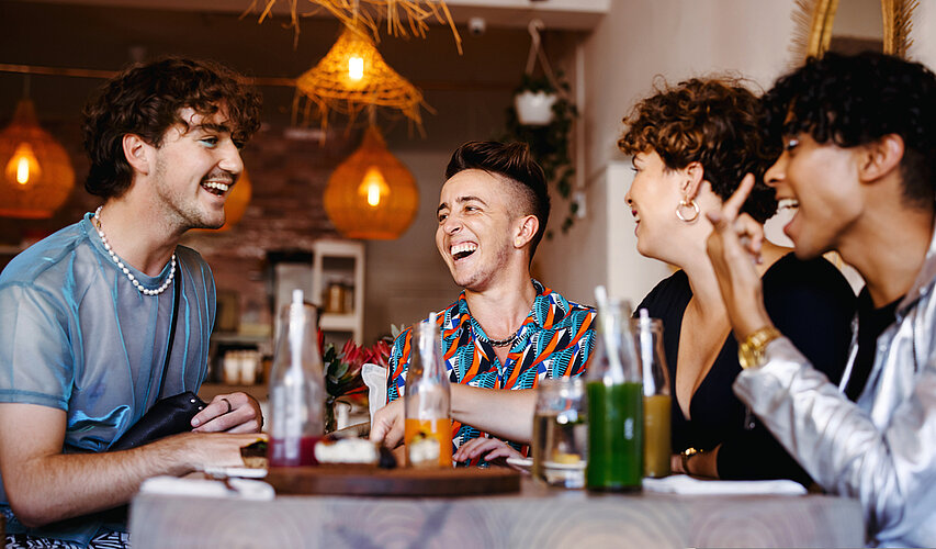 Vier Personen, die nicht eindeutig als Mann oder Frau zu lesen sind, sitzen fröhlich lachend an einem Tisch in einem Restaurant. Sie tragen verschiedenen Hemden, die bunt oder aus blauen Chiffon sind, sowie Perlenketten, Ohrringe und eine goldenen Uhr. Im Hintergrund hängen Lampenschirme aus Bast von der Decke. Auf dem Tisch stehen Flaschen mit verschiedenfarbigen Flüssigkeiten darin.