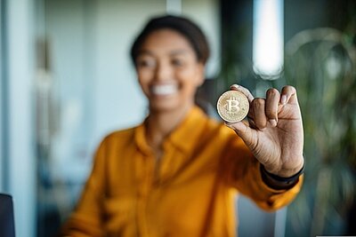 Bitcoin – Potenzial für finanzielle Chancengleichheit? Eine Frau hält eine Bitcoin-Münze in die Kamera. Die Frau ist unscharf im Hintergrund, die Münze in ihrer Hand deutlich und klar erkennbar.
