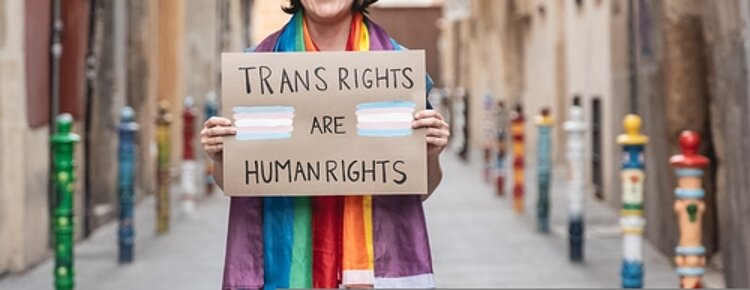 Ein weiterer Meilenstein: das Selbstbestimmungsgesetz In einer schmalen Gasse steht eine Person, die ein Transparent in der Hand hält mit der Aufschrift: Transrights are human rights. Die Person hat einen regenbogenfarbigen Schal um den Hals.
