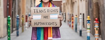 Ein weiterer Meilenstein: das Selbstbestimmungsgesetz In einer schmalen Gasse steht eine Person, die ein Transparent in der Hand hält mit der Aufschrift: Transrights are human rights. Die Person hat einen regenbogenfarbigen Schal um den Hals.