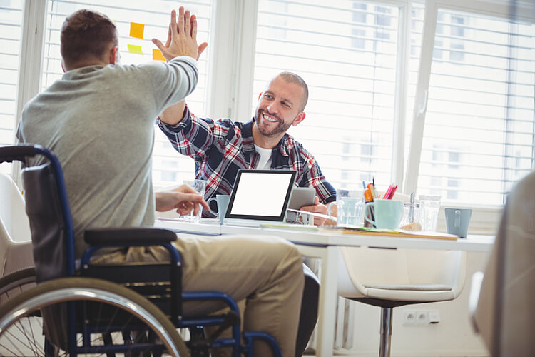 An einem Besprechungstisch geben sich zwei Männer eine High Five. Einer von beiden sitzt im Rollstuhl.