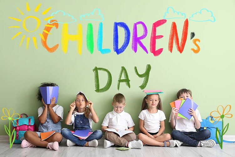 Fünf Kinder sitzen lachend oder spielerisch nebeneinander auf dem Boden vor einer hellgrünen Wand. Über ihnen steht bunt und kindlich gezeichnet ‚Children’s Day‘ mit Sonne, Wolken und Blumen. Die Kinder halten Bücher in verschiedenen Farben und wirken fröhlich und entspannt.